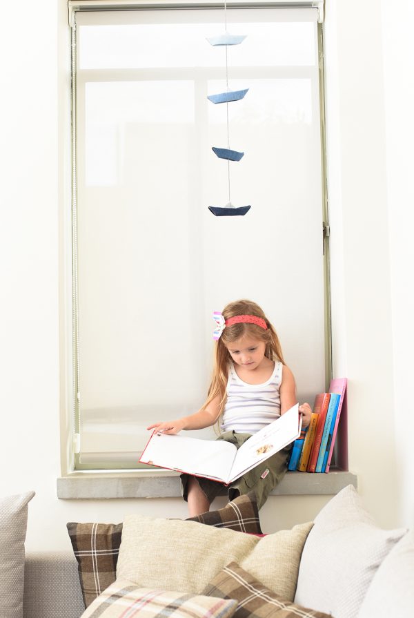 ילדה קוראת ספר עם סרט פרפר על הראש ומאחוריה גרלנדת סירות אוריגמי מבד ג'ינס כחול לעיצוב הבית.