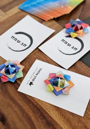 סביבון אוריגמי על גבי גלויית חג שמח עם סמל האנסו. סביבון ייחודי המקופל מניירות אוריגמי צבעוניים ויפהפיים , כל סביבון ייחודי ושונה. סביבון ממתוג, סביבון נייר, גלויית חג שמח, סדנאות סביבונים, סדנאות חנוכה, מתנות לחנוכה. מורן אלחלל